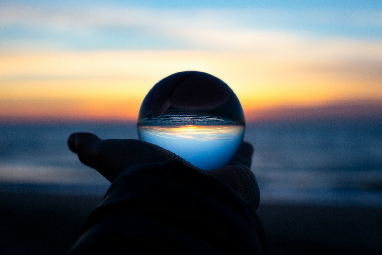 A hand holding a glass sphere reflecting a beach at low sun